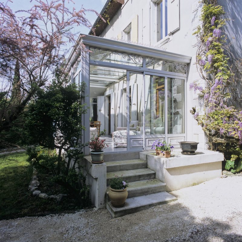 Véranda art déco blanche et lumineuse, en aluminium, adossée à la façade d’une maison.